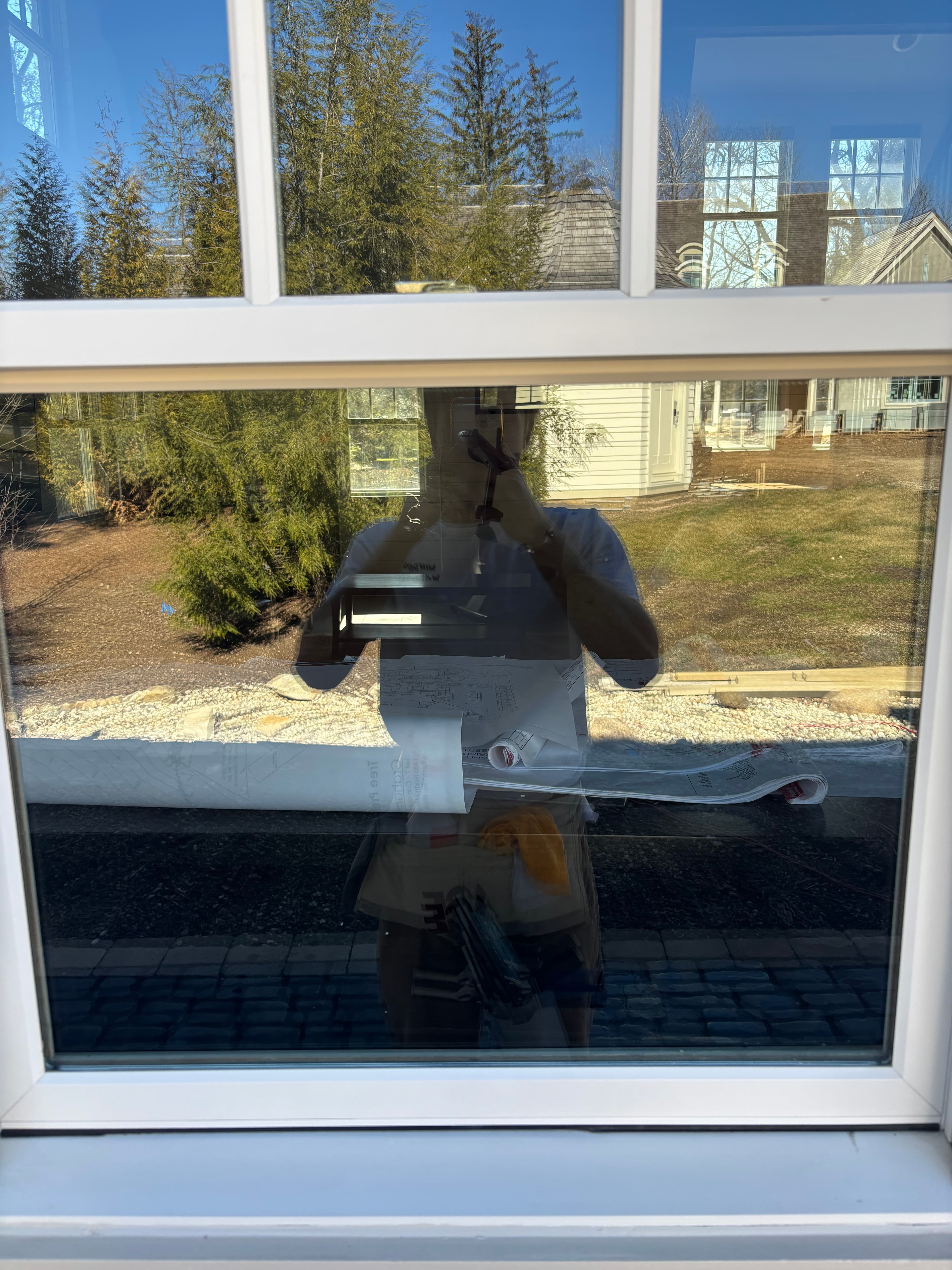 Reflection of a person holding blueprints in a window overlooking a yard with trees.